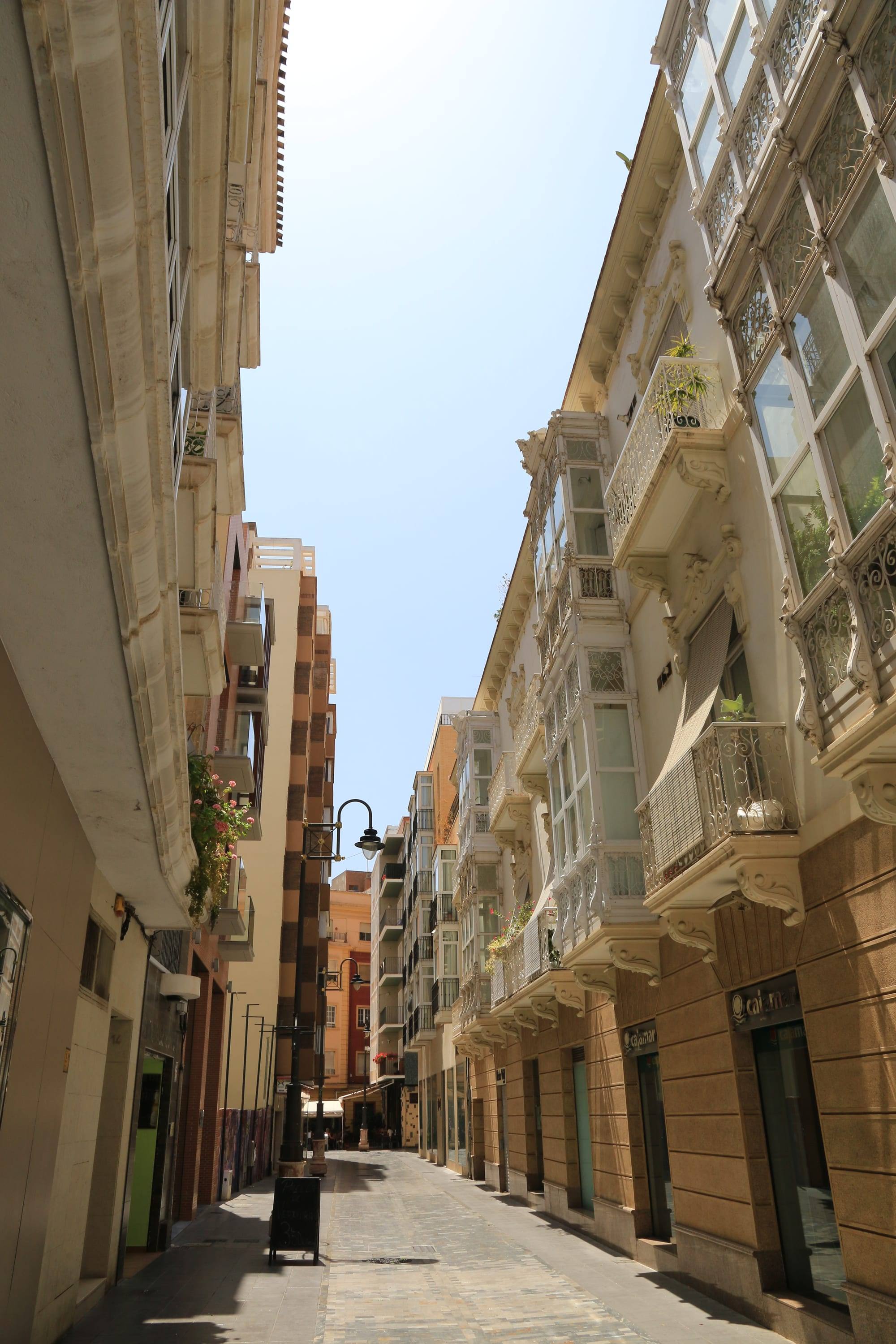 A narrow pedestrian street in Cartagena's old town seen from below: on the right, cream façades with ornate white glazed miradores, wrought-iron balconies and carved corbels catching the sun, on the left plain shaded walls, a black lamp post and a chalkboard sign on the paved lane, and a strip of pale blue sky between the buildings