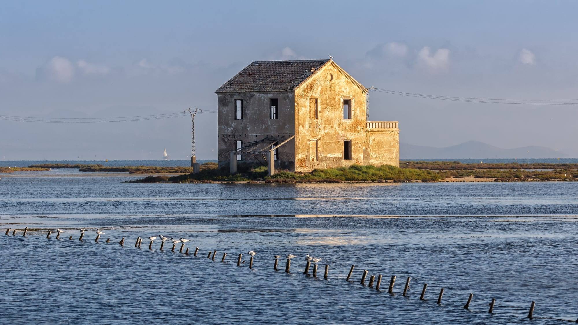 An abandoned two-storey fishermen's house of weathered ochre plaster with a tiled roof, standing on a grassy islet in the shallow water of the Mar Menor at Las Encañizadas, La Manga, glowing in low evening sun; a curving line of wooden stakes with perched seagulls crosses the rippled blue water in front, with a white sail and distant hills on the horizon