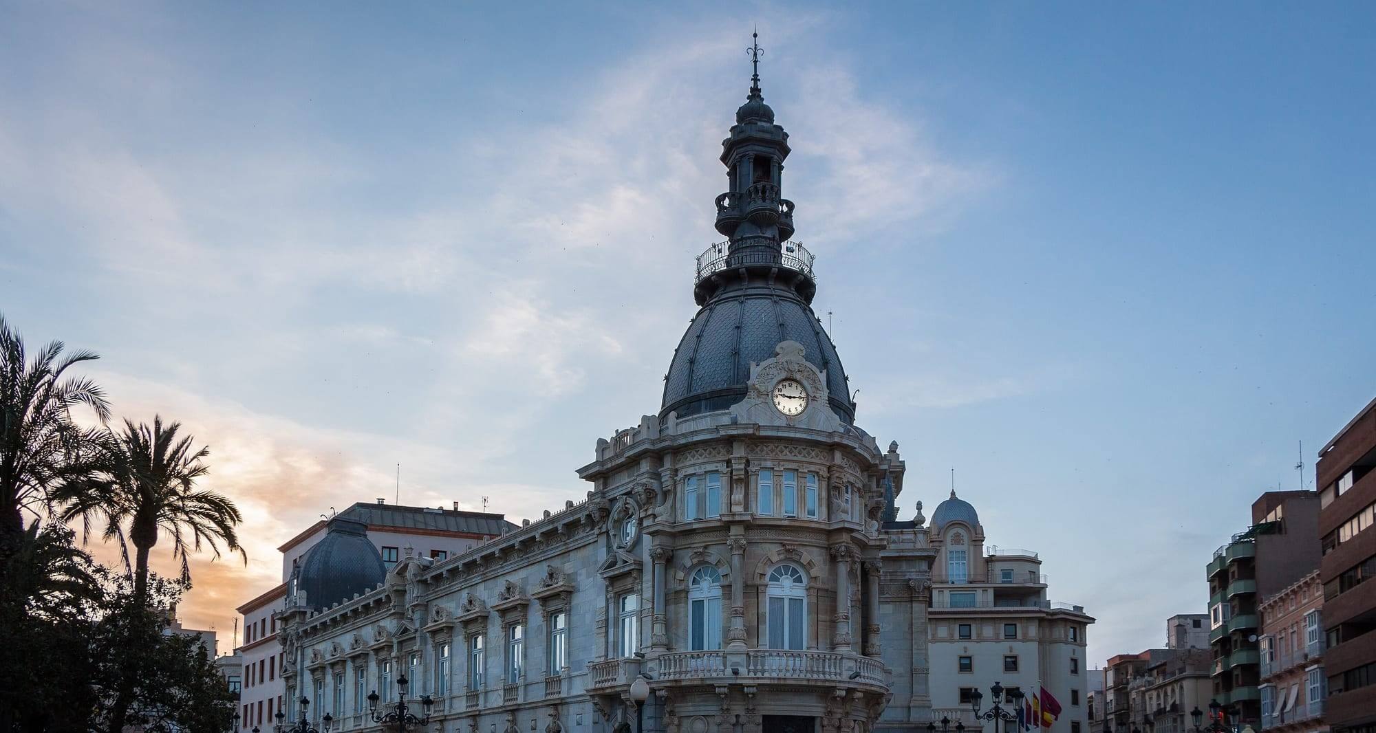 The Palacio Consistorial, Cartagena's modernista town hall, at dusk: the corner rotunda with its clock, slate dome and wrought-iron lantern, a long façade of pale stone and grey marble ending in a second dome, and palm trees silhouetted against a peach sunset on the left