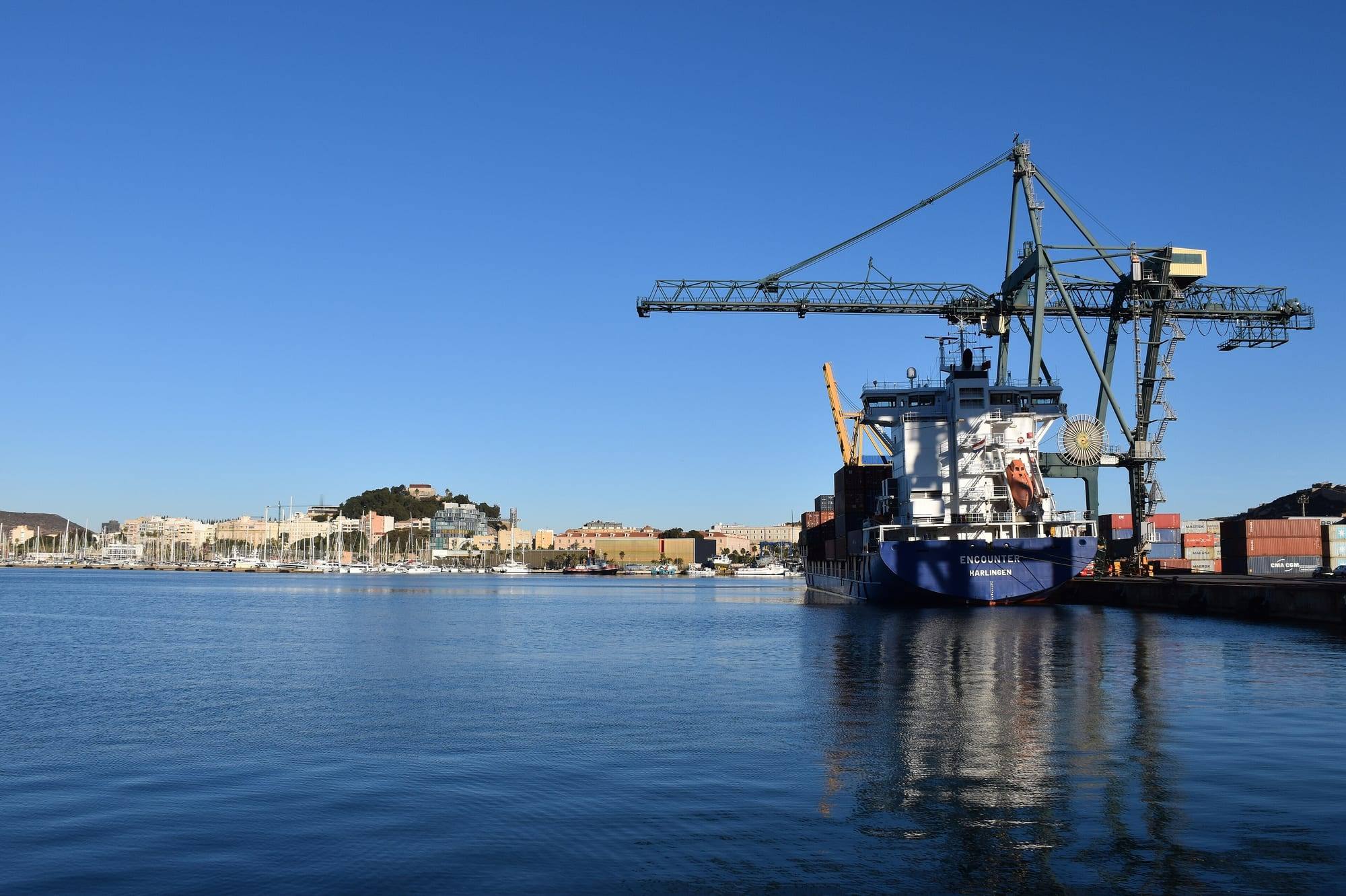 The commercial quay of the port of Cartagena on a clear winter morning: a dark-blue container ship moored under a tall grey-green gantry crane, stacked containers on the quay at the right, the calm deep-blue water of the harbour reflecting the hull, and across the basin the masts of the marina, the pale seafront buildings and the pine-covered hill of the Castillo de la Concepción under a cloudless sky