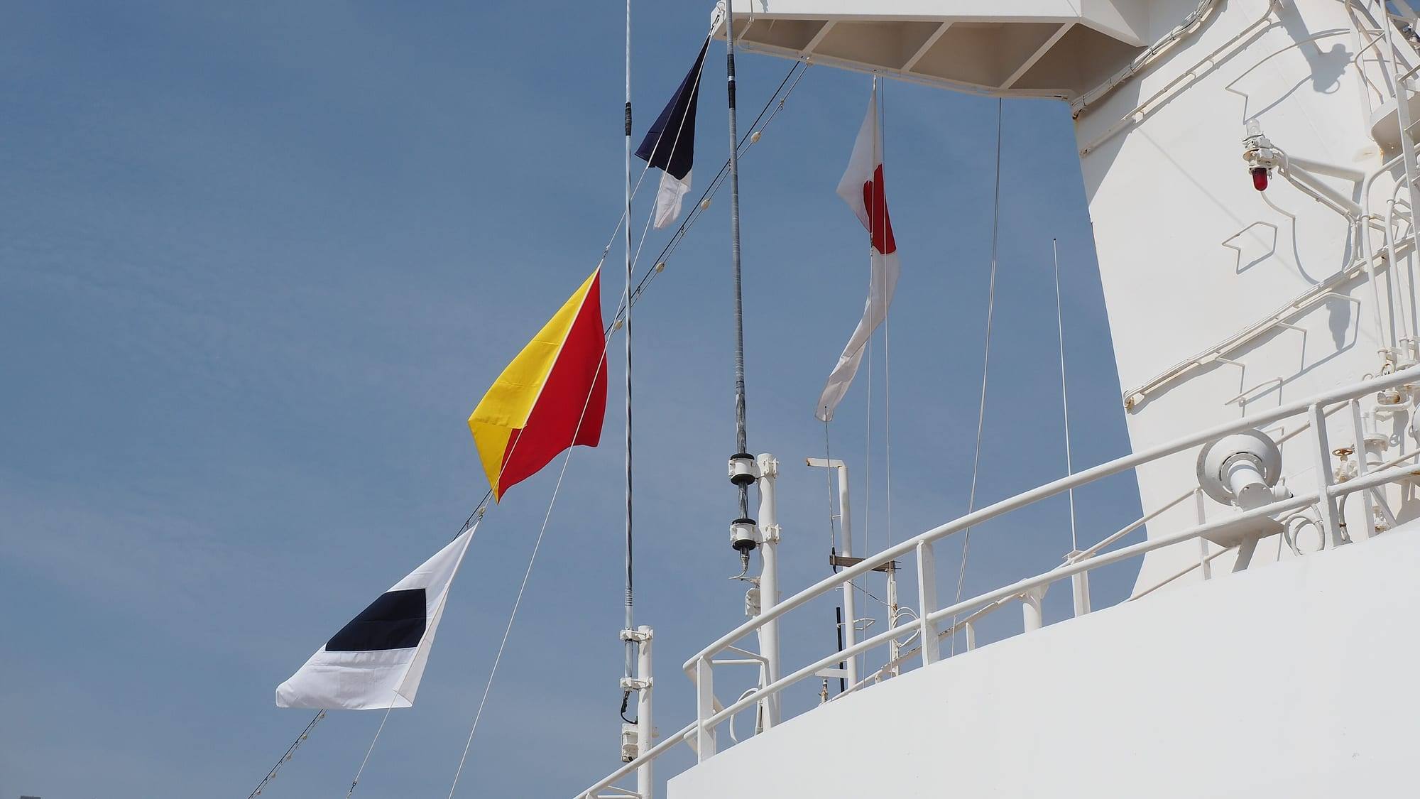 Signal flags hoisted on the mast of a ship entering the port of Yokohama, Japan: the dark-blue-and-white second substitute pennant above the diagonal red-and-yellow 'O' flag and the white 'S' flag with its dark-blue centre square, the Japanese courtesy flag flying beside them, against a pale blue sky and the white superstructure and railings of the ship